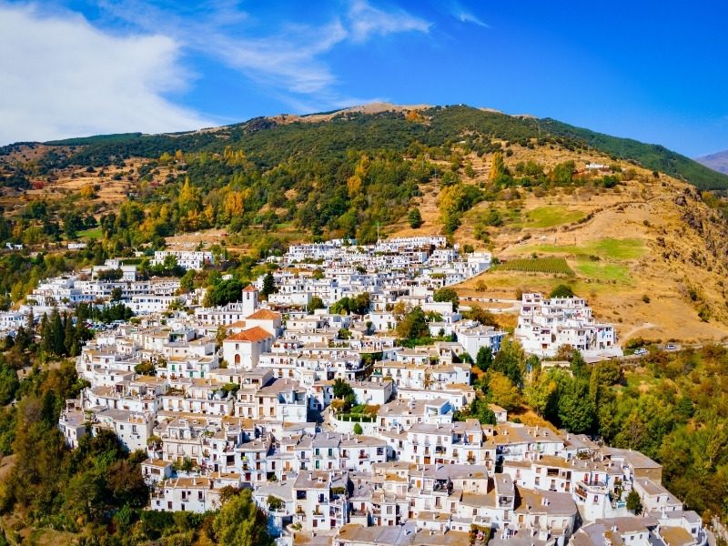 A whitewashed Alpujarras village with flat roofs stacked up a green hillside in fall, on a self-guided hiking tour in southern Spain