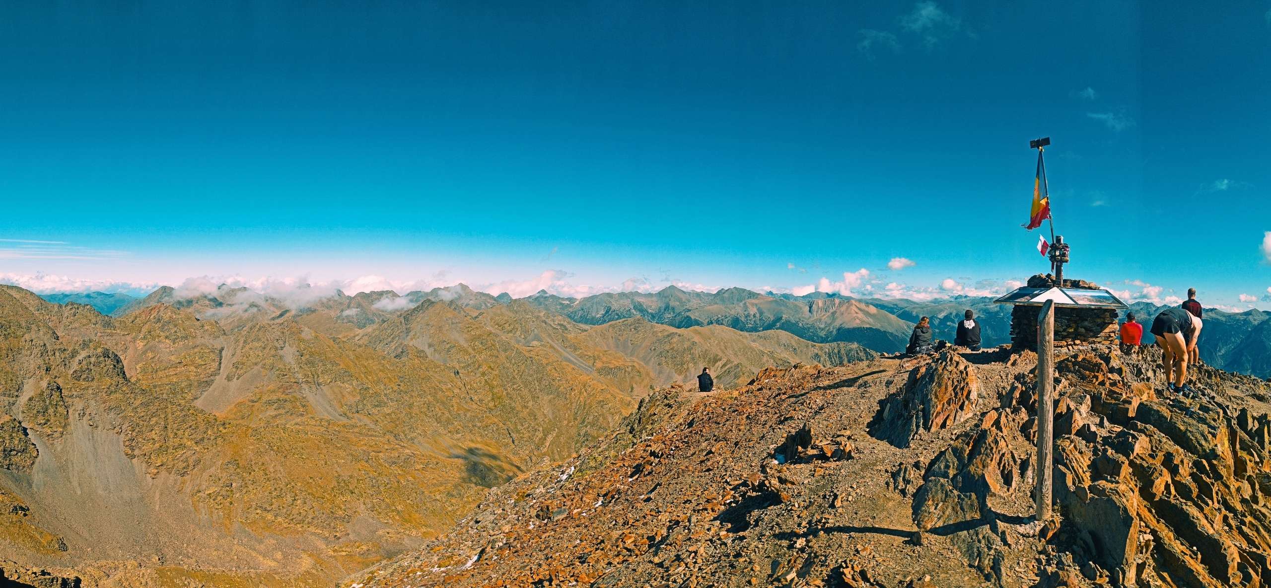 Hikers at the summit marker and the Andorran flag, looking out over range after range of the Pyrenees, on a self-guided Andorra hiking tour