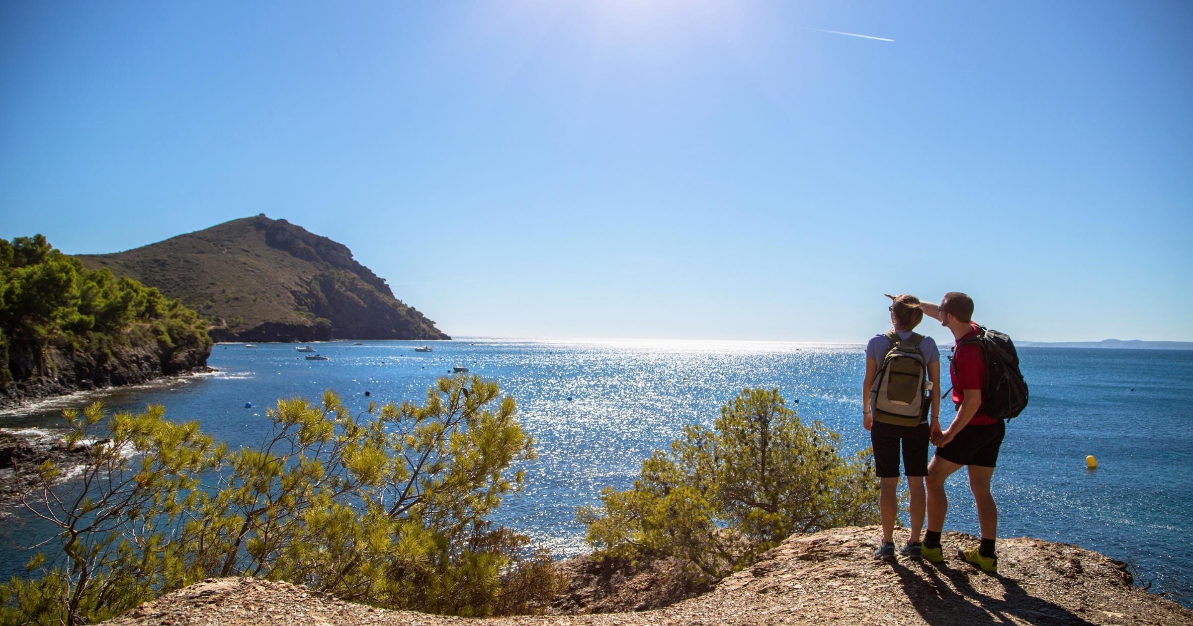 Two hikers looking out over a sparkling Mediterranean cove from a rocky viewpoint on a self-guided Costa Brava hiking tour in Catalonia, Spain
