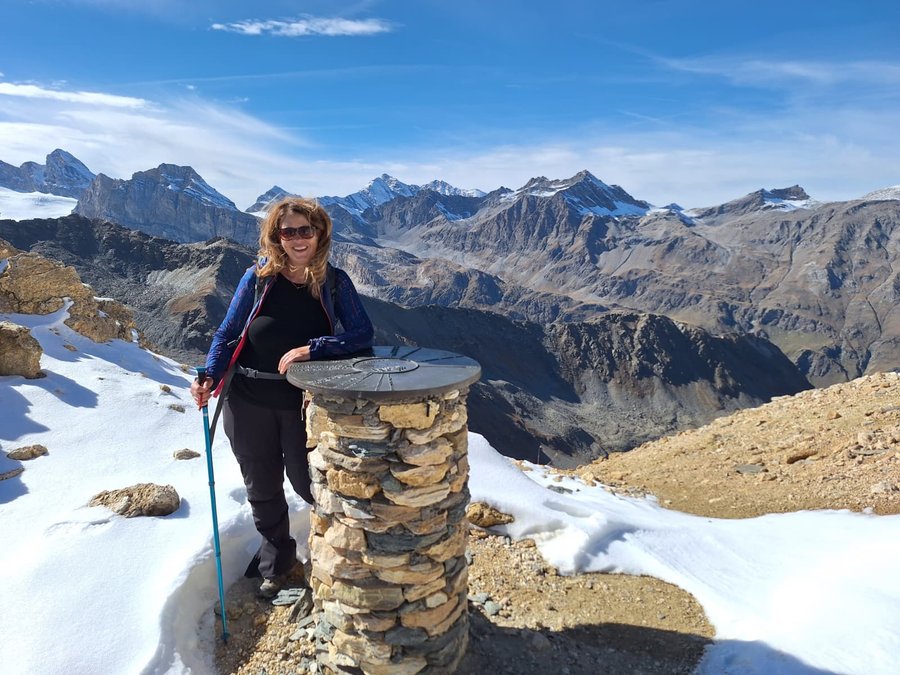 A hiker beside the stone summit marker at the Col Rosset, with the peaks of Gran Paradiso National Park behind