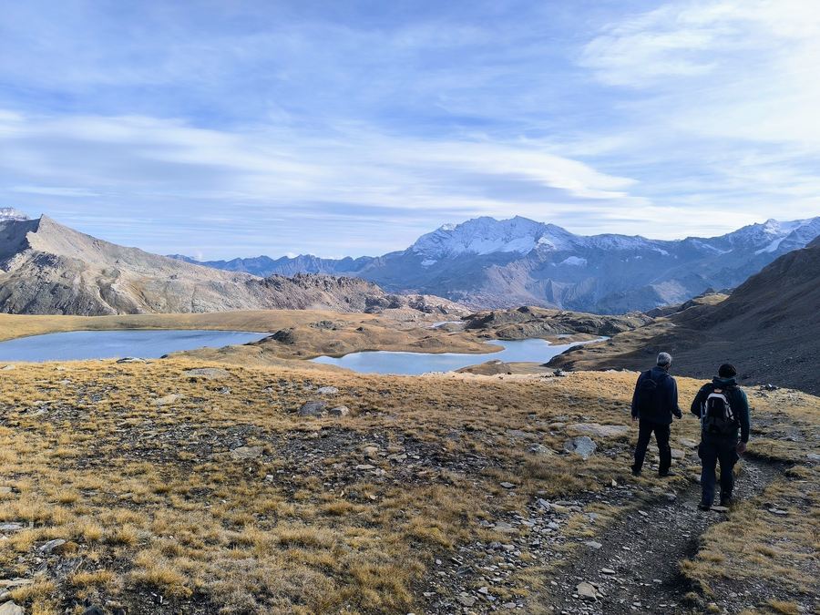 Two hikers descending a grassy trail towards the lakes of the Plan del Nivolet in the Italian Alps