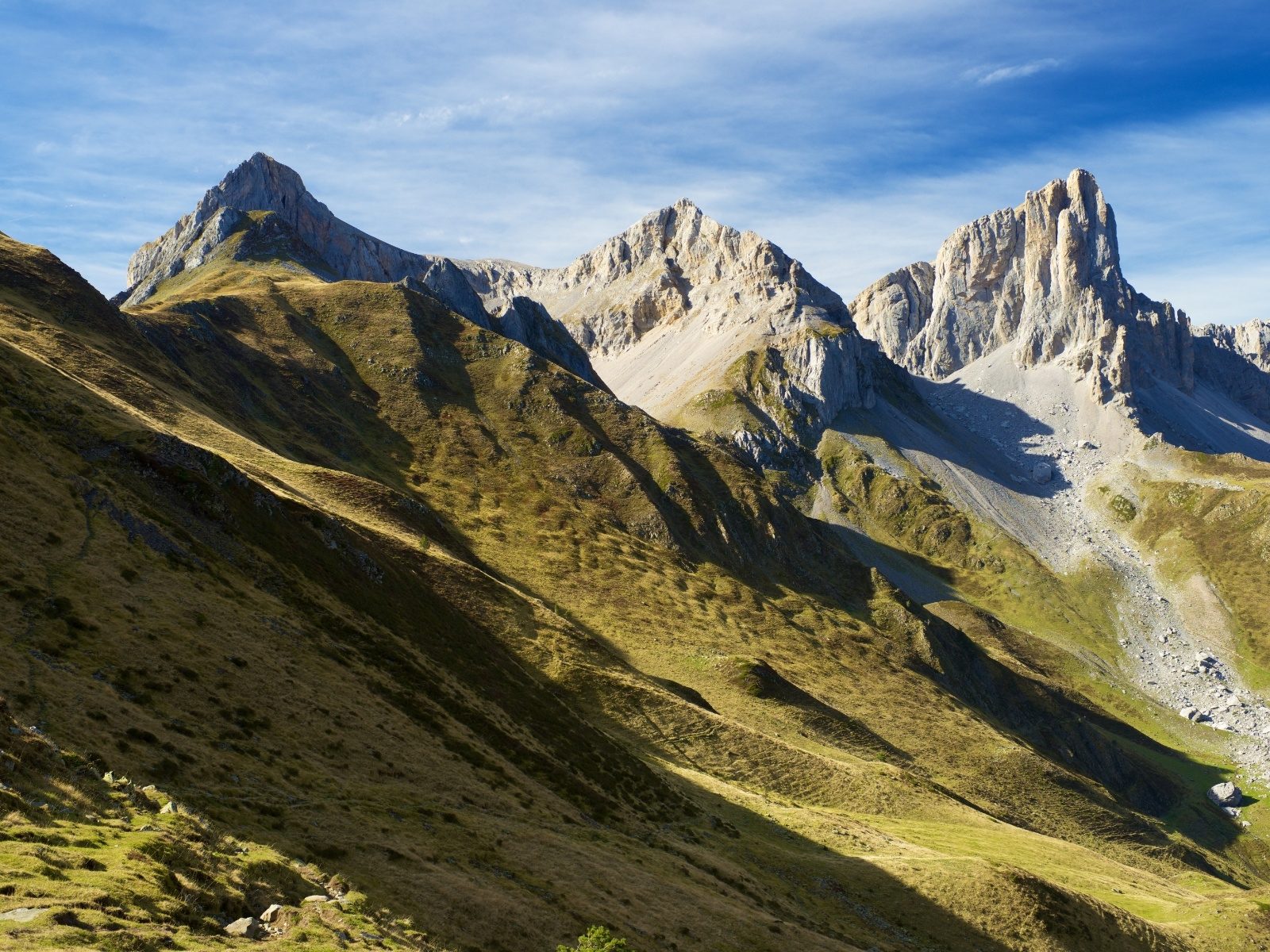 Jagged limestone peaks above a green ridge with a hiking trail traversing it in the Pyrenees