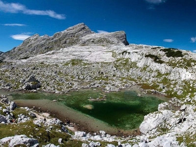 A small green tarn held in bare grey limestone below the peaks of the Julian Alps on the hut to hut route