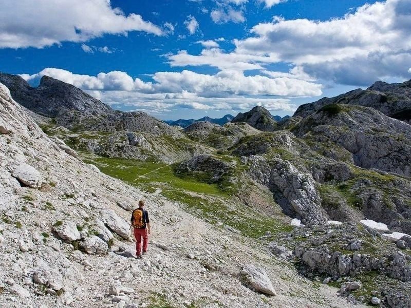 A hiker in a red jacket on a stony trail crossing an open limestone basin in Triglav National Park on the hut to hut route