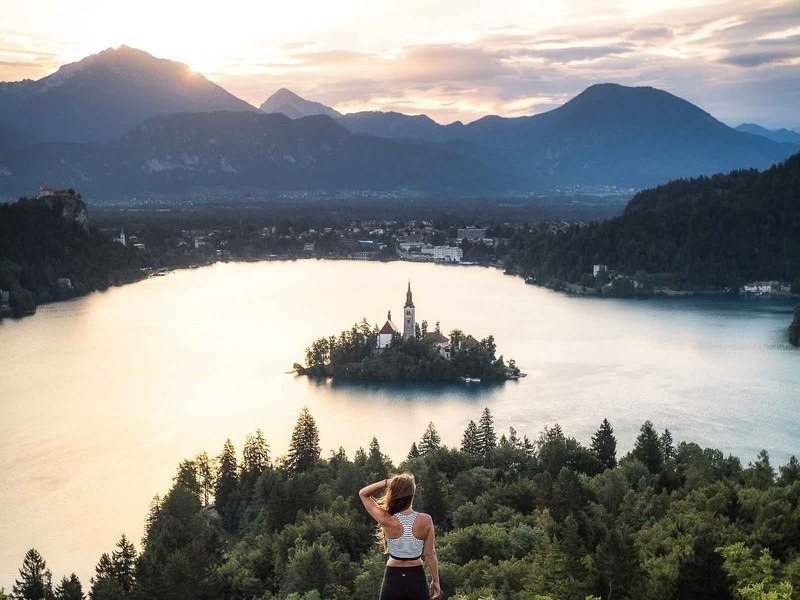 A hiker with arms raised above Lake Bled at sunrise, with the island church below and Bled castle on its cliff