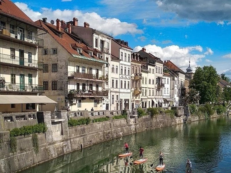 Pastel townhouses and cafe terraces along the Ljubljanica river in Ljubljana, with paddleboarders on the water