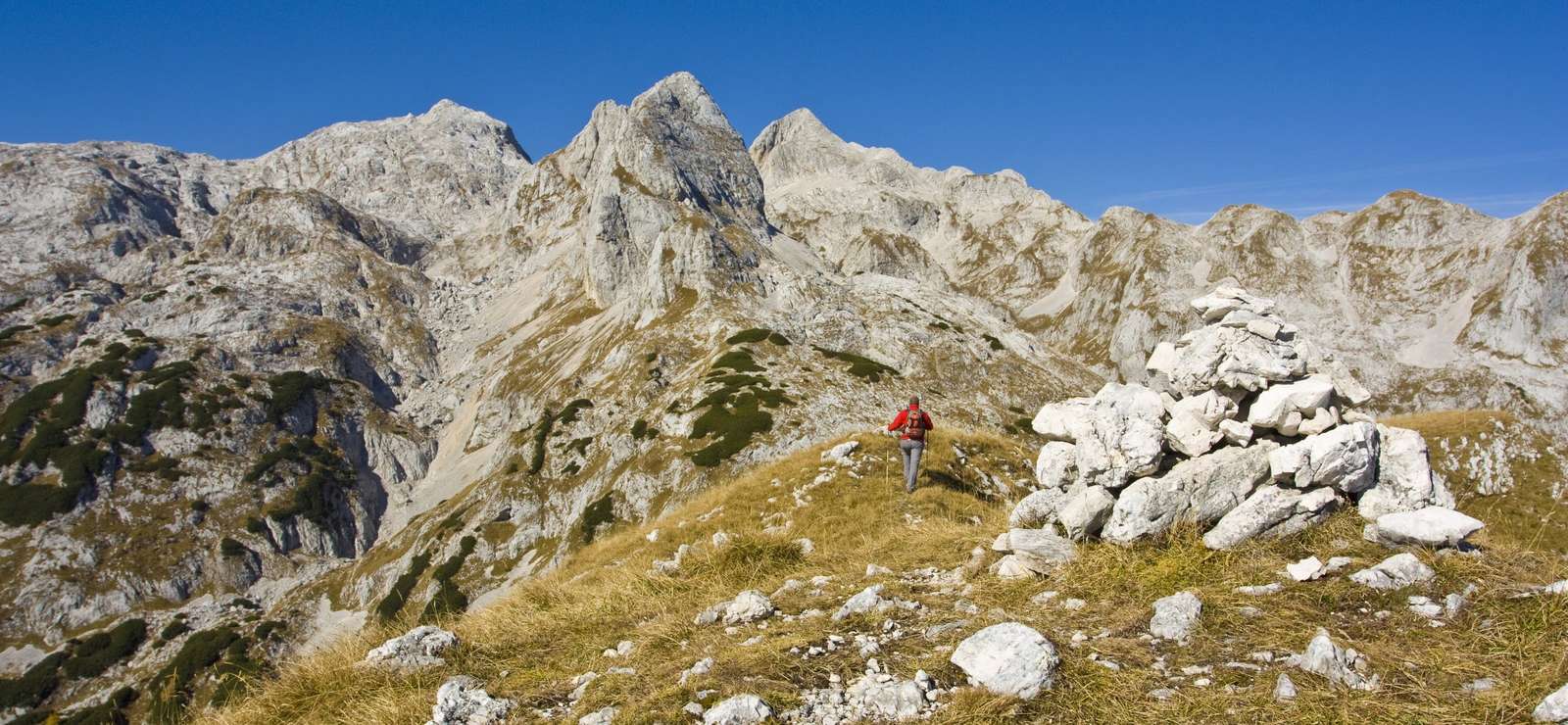 A hiker on a grassy ridge beside a limestone cairn below the jagged peaks of the Julian Alps on our Slovenia hiking tour