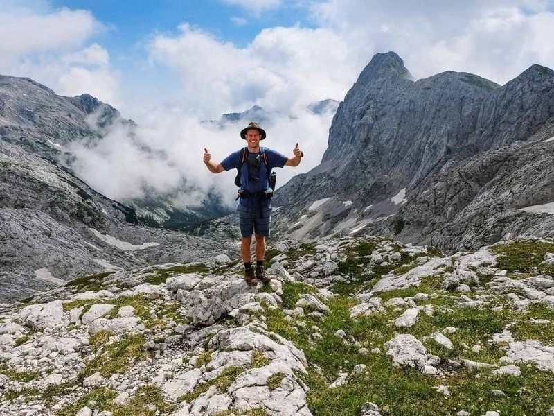 A hiker standing with arms out on rocky ground high in the Julian Alps with peaks all around, on the Slovenia hut to hut hike